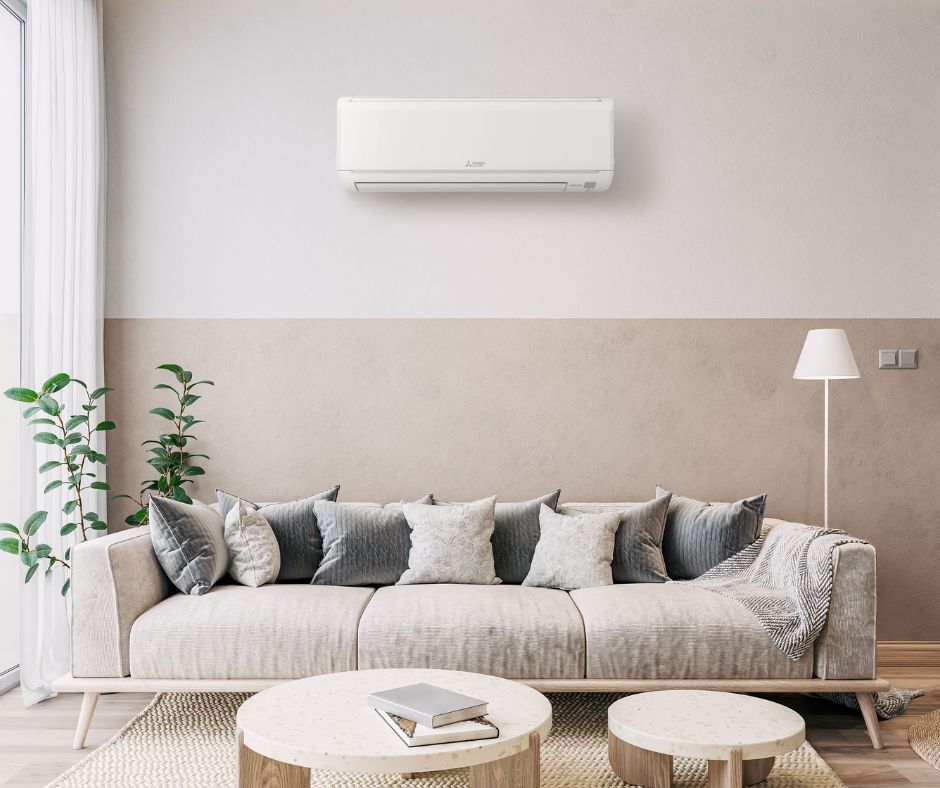 Aire condicionat tipus split instal·lat a la paret sobre un sofà en un saló modern, amb decoració minimalista en tons neutres, plantes decoratives i llum natural entrant des d’un balcó.